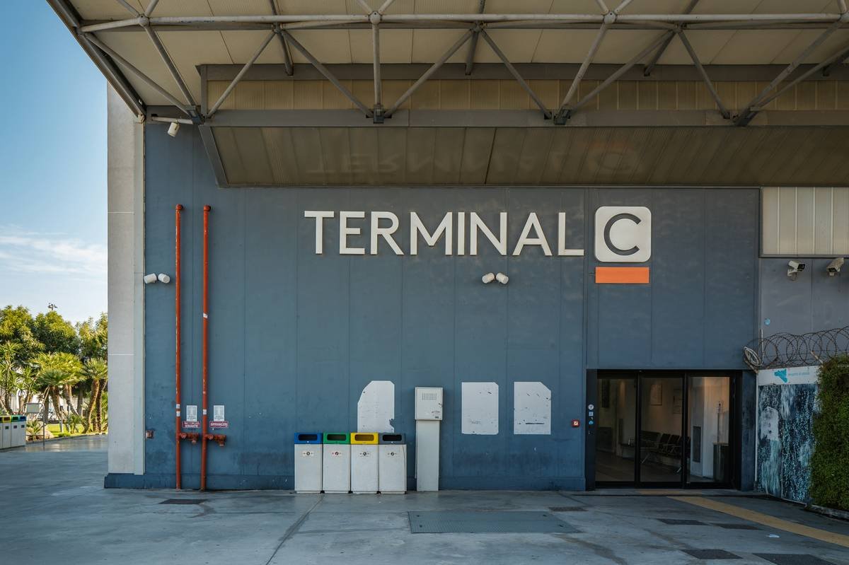 This photograph captures a modern terminal building at Catania Airport in Italy. The focus is on the 'TERMINAL C' signage, prominently displayed in white against a vibrant blue facade. The building's architecture is characterized by clean lines and a flat roof, indicative of contemporary airport design. Several security cameras are mounted on the building, emphasizing the focus on safety and security. The surrounding area includes concrete pavement, trash receptacles, and a fire hydrant, contributing to the functional and utilitarian atmosphere of an airport environment.